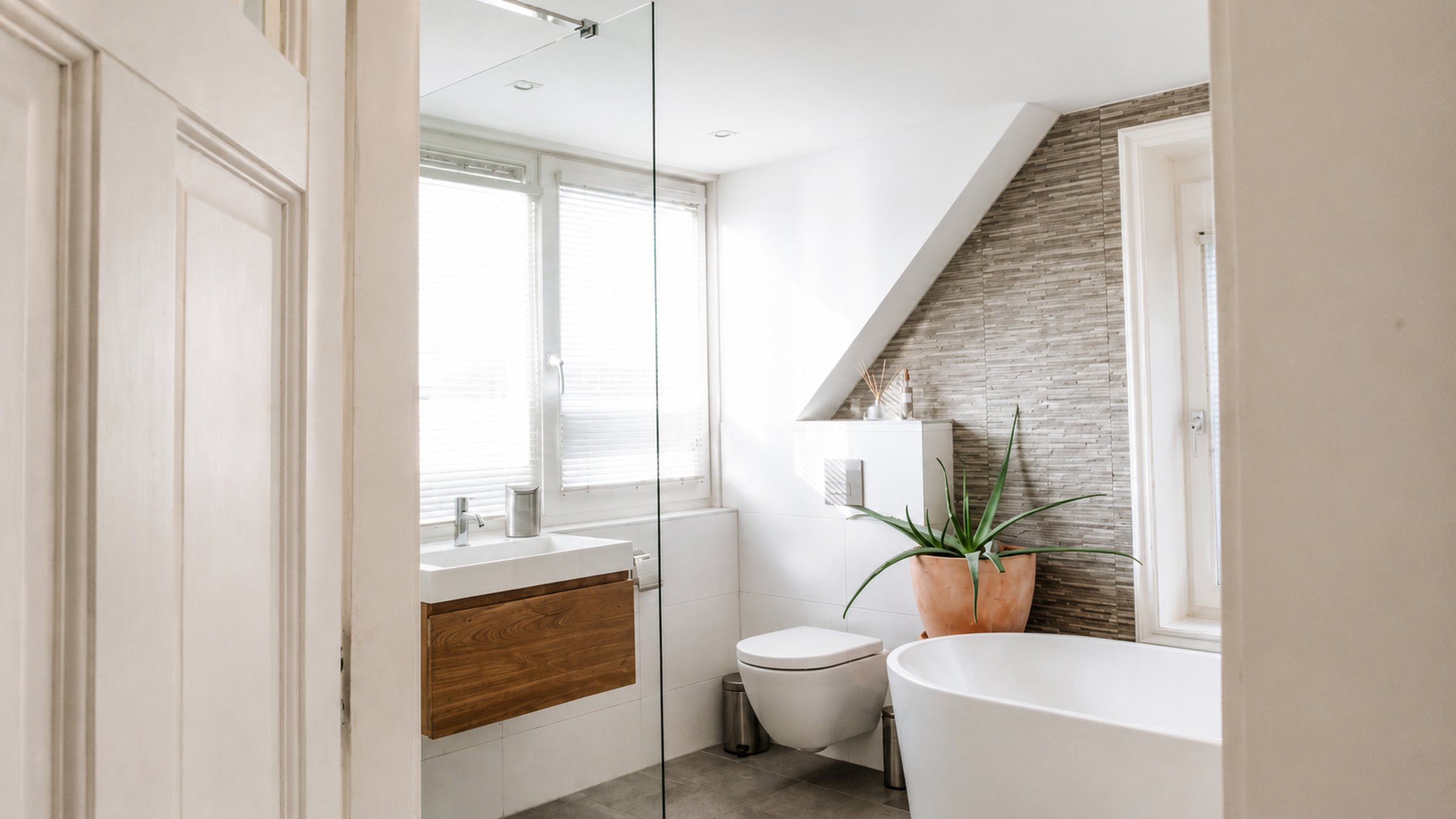 The renovated Schoorl bathroom seen from the doorway, with glass shower partition, freestanding bath, natural stone tiles and light from two sides