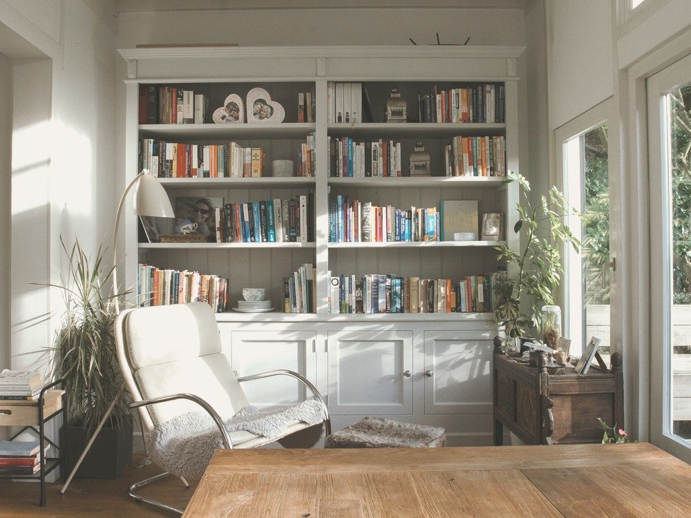 A sun-filled reading nook with floor-to-ceiling built-in bookshelves painted in warm grey, a white leather reclining chair, and large windows overlooking the garden