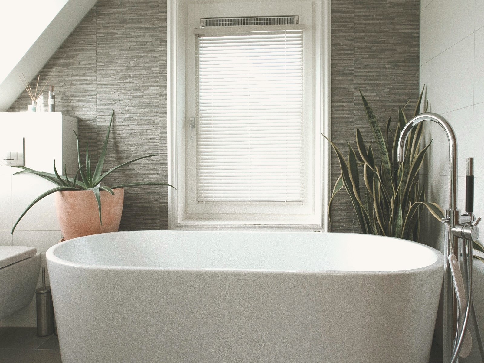 A serene attic bathroom with a freestanding white bath flanked by two large plants, full-width window with view towards the Schoorlse Duinen, and grey natural stone wall tiles