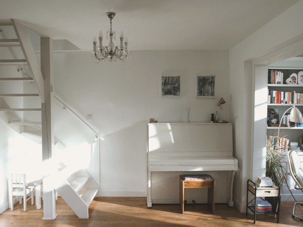 A bright entrance hall with a white upright piano, crystal chandelier, dappled natural light across white walls, and a glimpse of the bookshelf room to the right