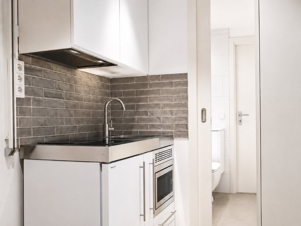 Compact kitchenette with grey tile backsplash, stainless steel counter and bathroom beyond, micro apartment De Pijp Amsterdam - Ruimte interior design
