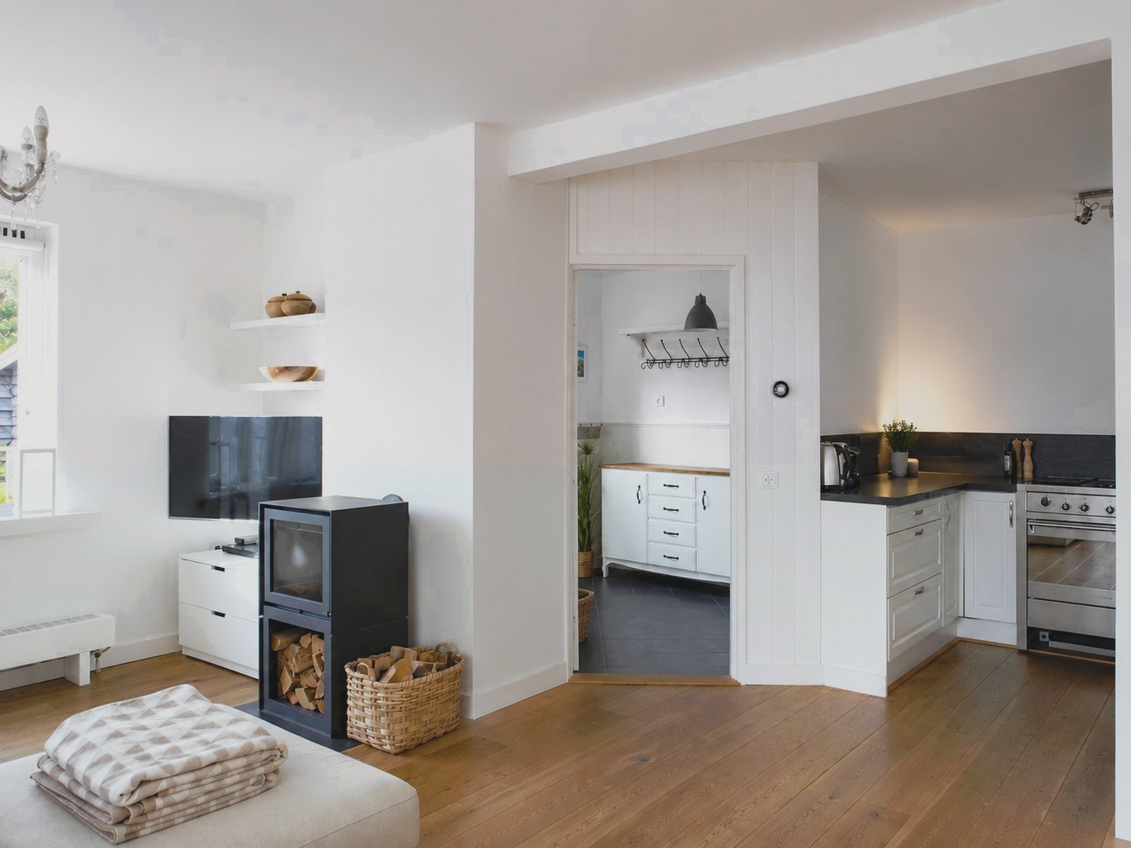 Open plan living area in the renovated Schoorl family home showing a black wood-burning stove, white kitchen with dark worktop, and warm oak floors throughout
