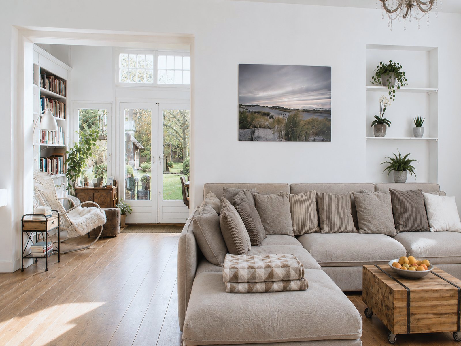 A bright open living room in the renovated Schoorl family home, with wide oak floors, large sectional sofa, French doors opening to the garden, a dunes canvas print and a chandelier above